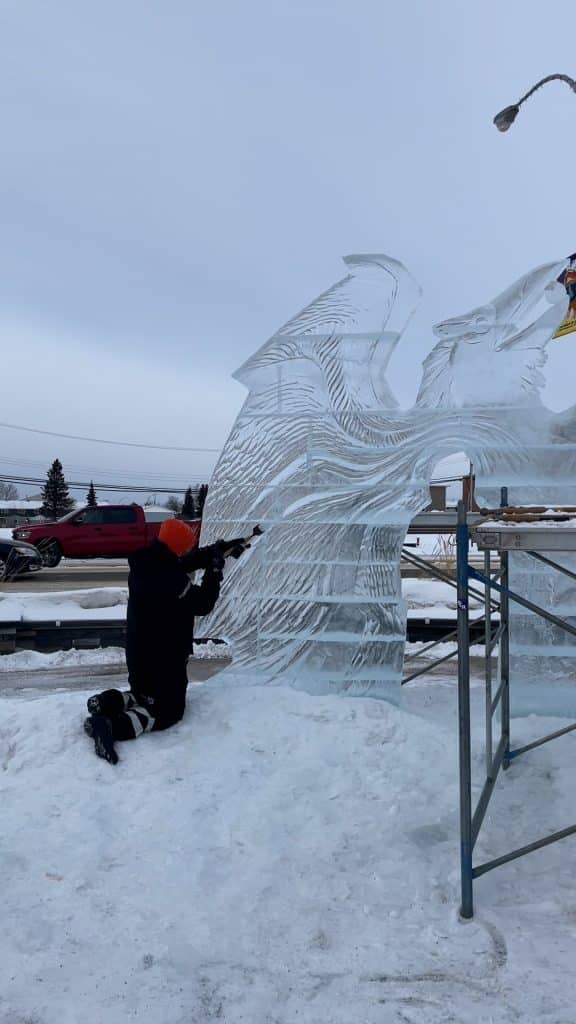 A man working on an ice sculpture with a truck driving behind the sculpture.