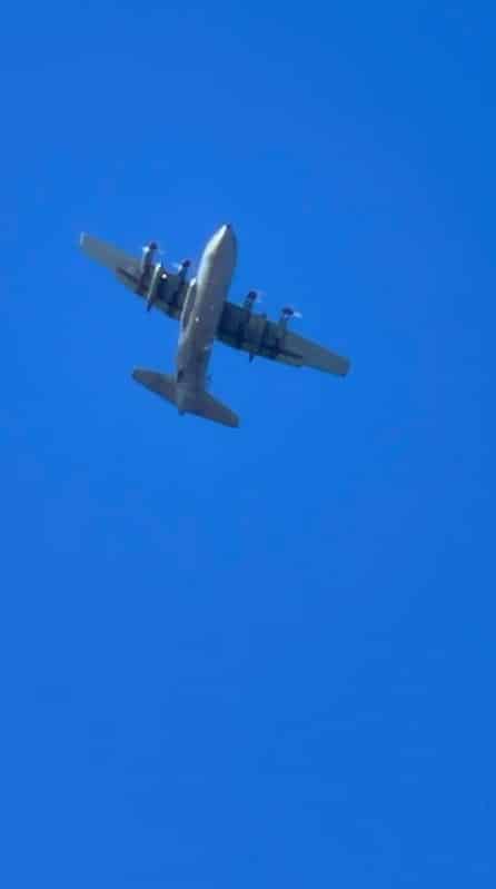 A grey plane with four propellors on the front flying in a bright blue sky.