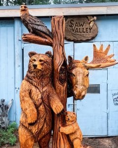 A Ryan Cook wood carving in front of a wooden sing that says Saw Valley.