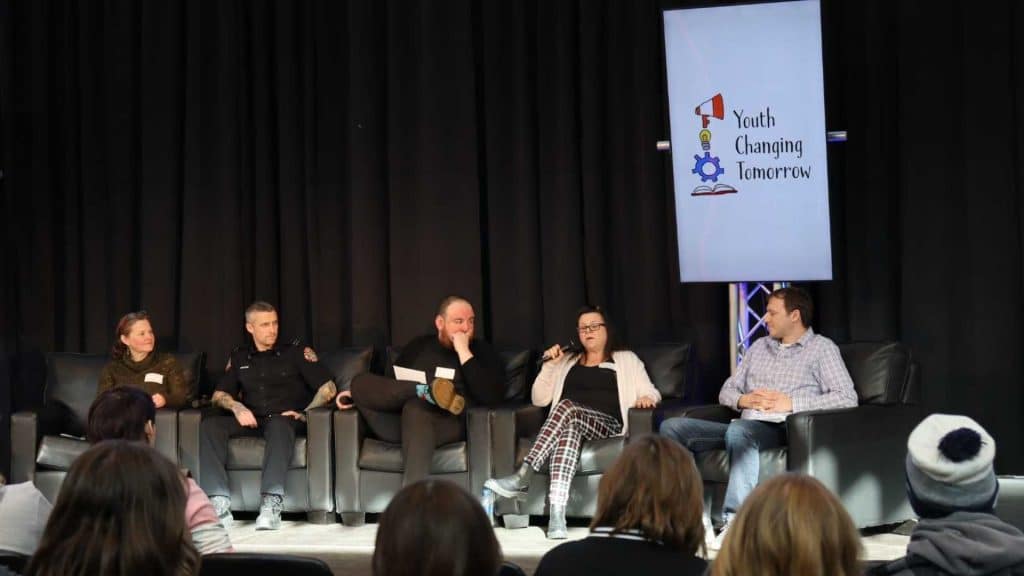 Five people sit on a stage, panel style. A sign next to them reads 'Youth Changing Tomorrow'