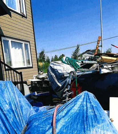 A photo of a lot of junk and furniture, and something covered by a blue tarp, in front of a house.