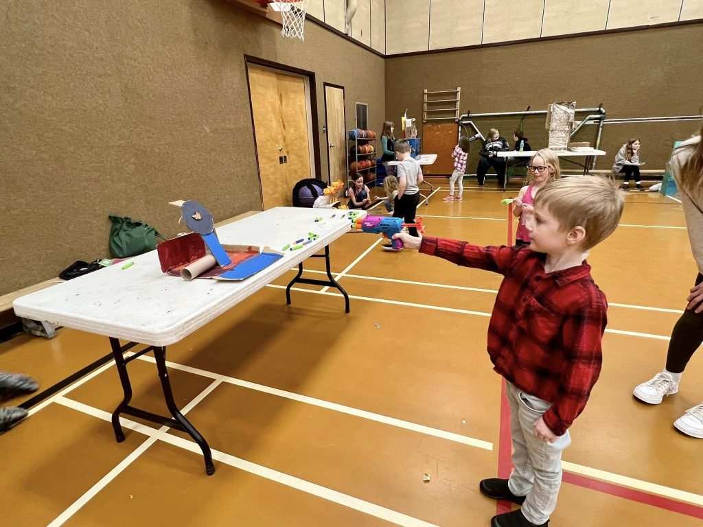 A student holding a nerf gun at a cardboard target in a school gym with other students around him.