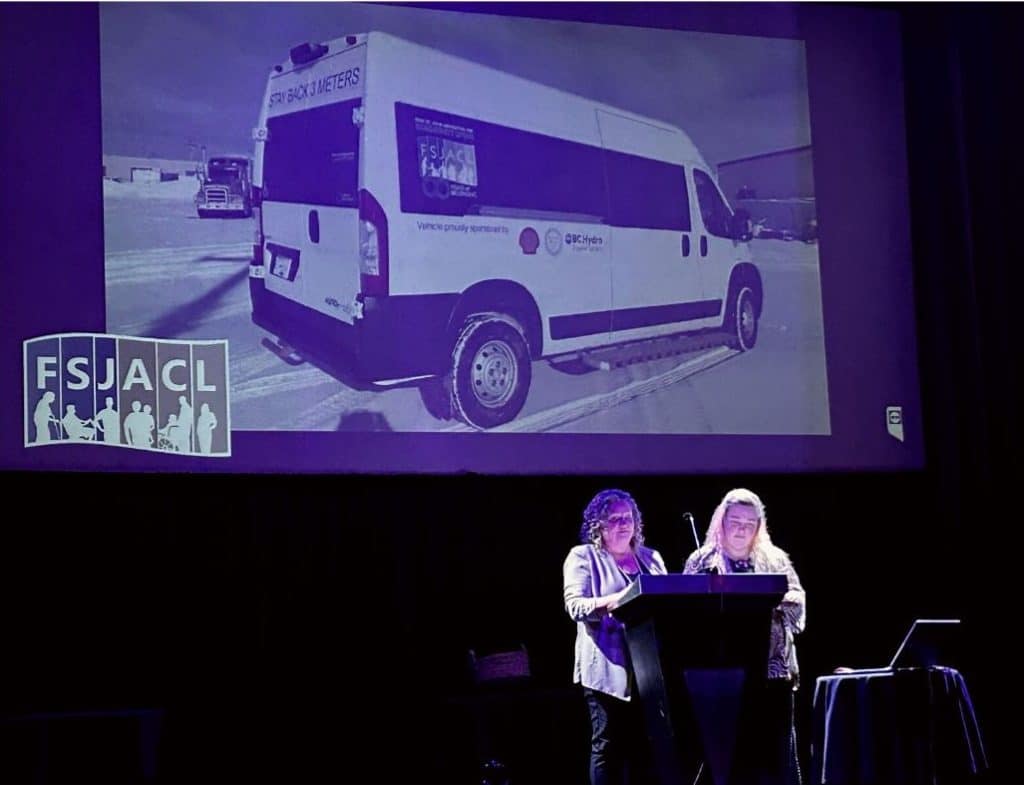 Two women standing behind a podium in front of a screen with a van displayed on it.
