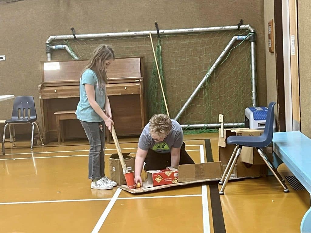Students setting up and playing a cardboard arcade game in a school gym.