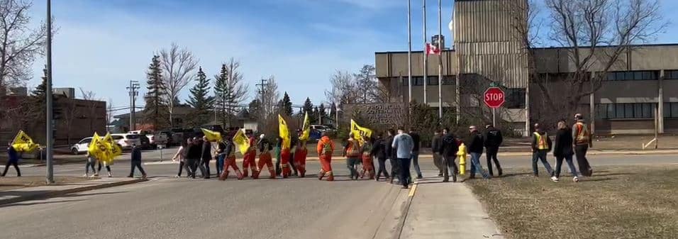 A group of people, some wearing high visibility vests and jackets, walking across the street. Some are holding yellow flags.