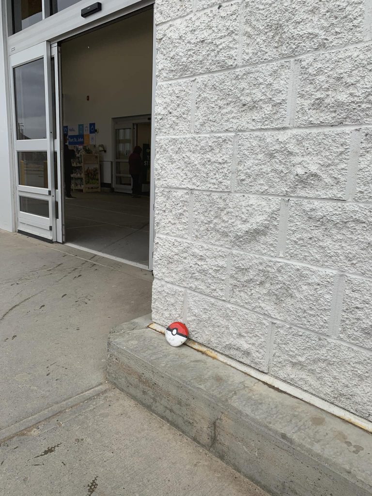 A rock painted like a pokeball sitting against a grey brick building next to automatic doors.