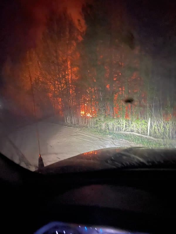 A photo taken from inside of a vehicle of a wildfire just behind some trees.