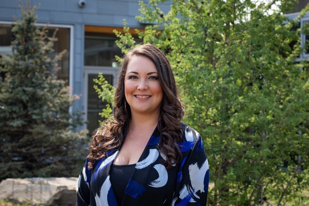 A smiling woman standing in front of a building and some evergreen trees.